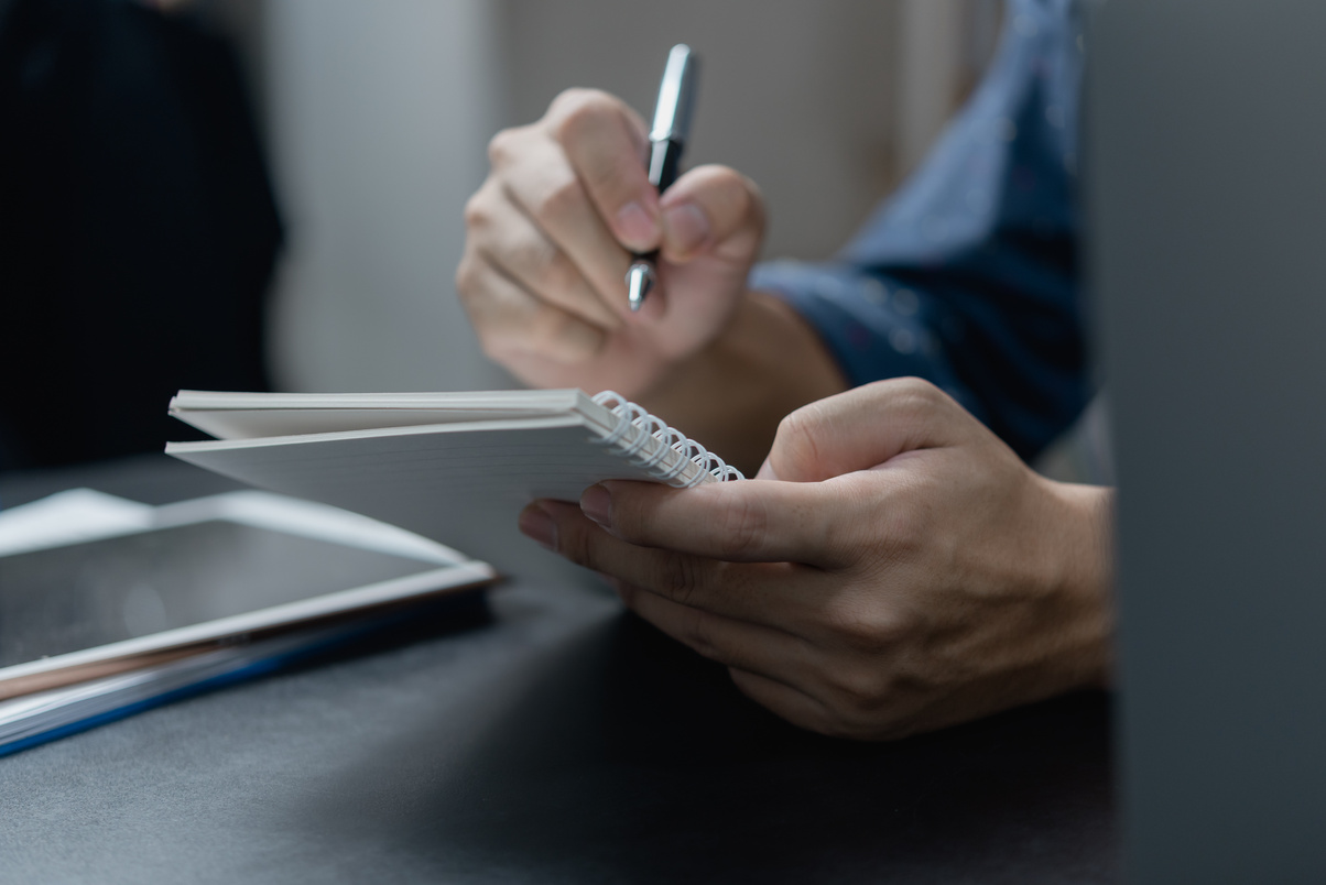 male hand writing in notebook with pen on laptop computer in modern office.